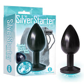 The image showcases "The Silver Starter Butt Plug," a beginner-sized plug measuring 2.8 inches, featuring an anodized silver and aqua finish. The packaging highlights its jeweled base with a close-up of the blue jewel and includes a black velvet pouch with a drawstring, all displayed elegantly beside the box.